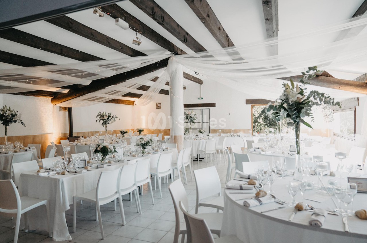 Salle de réception lumineuse avec tables décorées de nappes blanches, vaisselle élégante et arrangements floraux.