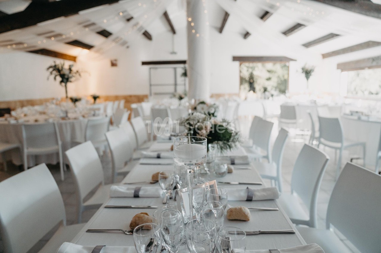 Salle de réception lumineuse avec tables dressées, nappes blanches, décorations florales et chaises blanches.