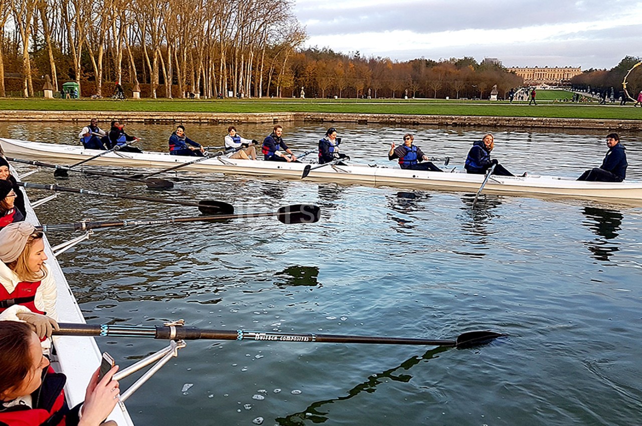 Des personnes rament sur des barques dans un plan d'eau entouré d'arbres, avec un bâtiment en arrière-plan.