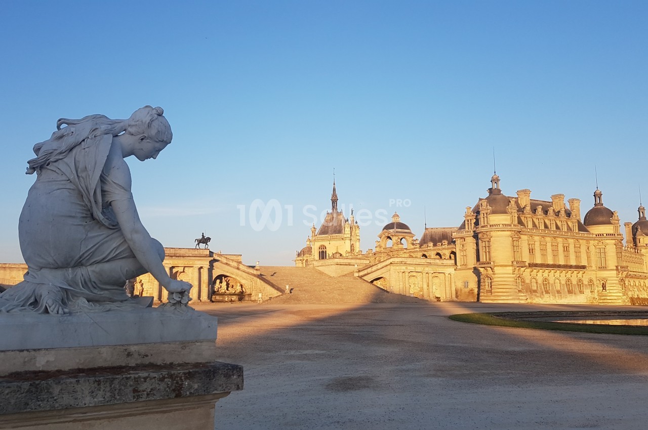 Statue en marbre au premier plan avec le château de Chantilly et son pont illuminés par le soleil couchant en arrière-plan.