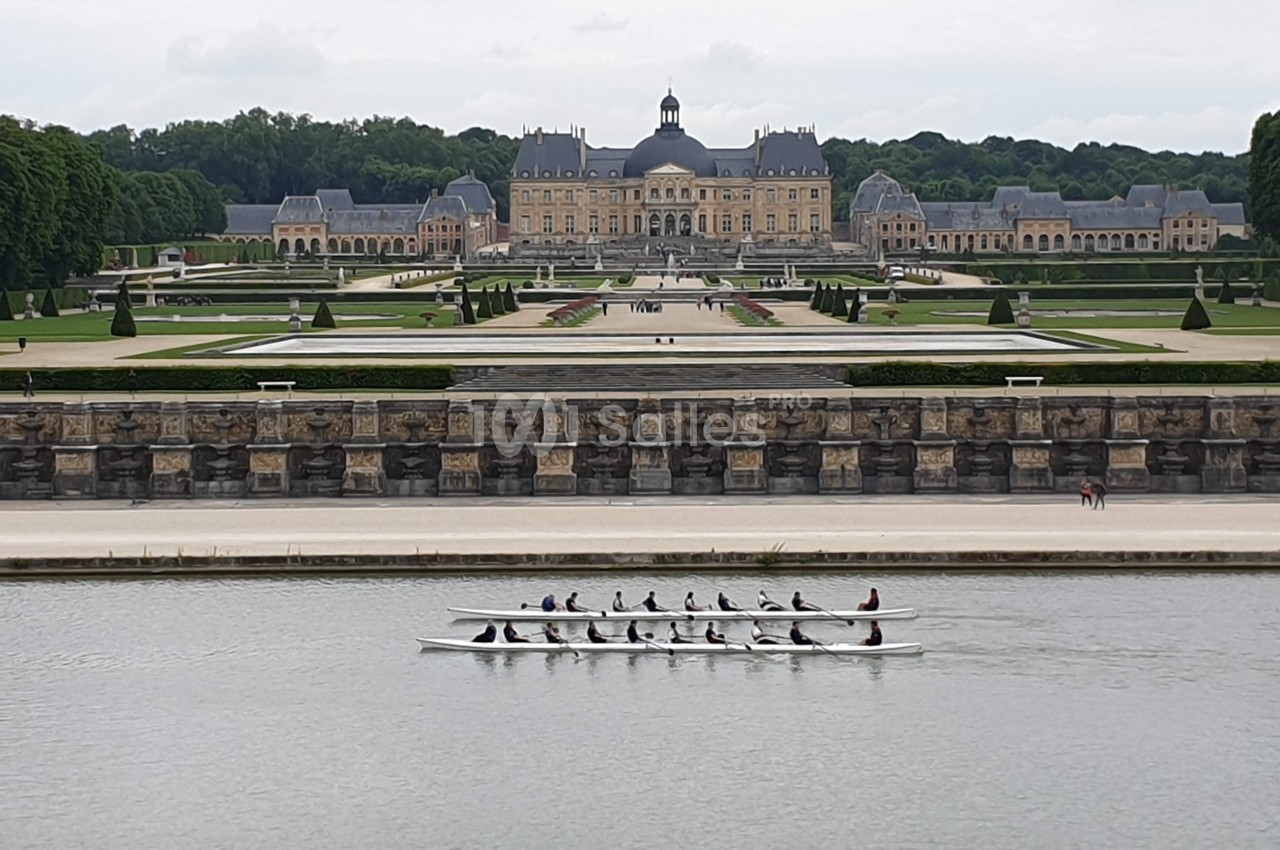 Un groupe de personnes rame dans une embarcation sur un canal, avec un château et ses jardins en arrière-plan.