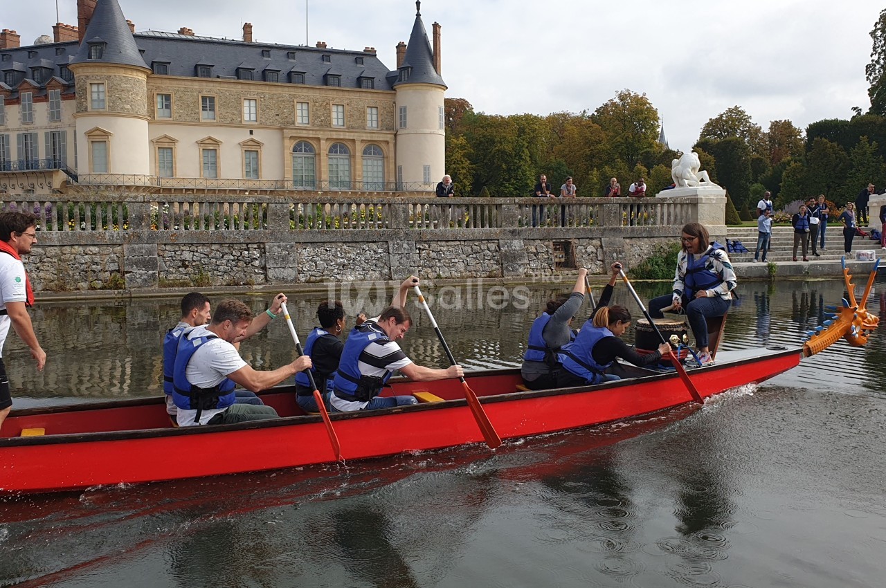 Des personnes pagaient dans une grande barque rouge sur un plan d'eau, près d'un château entouré de verdure.