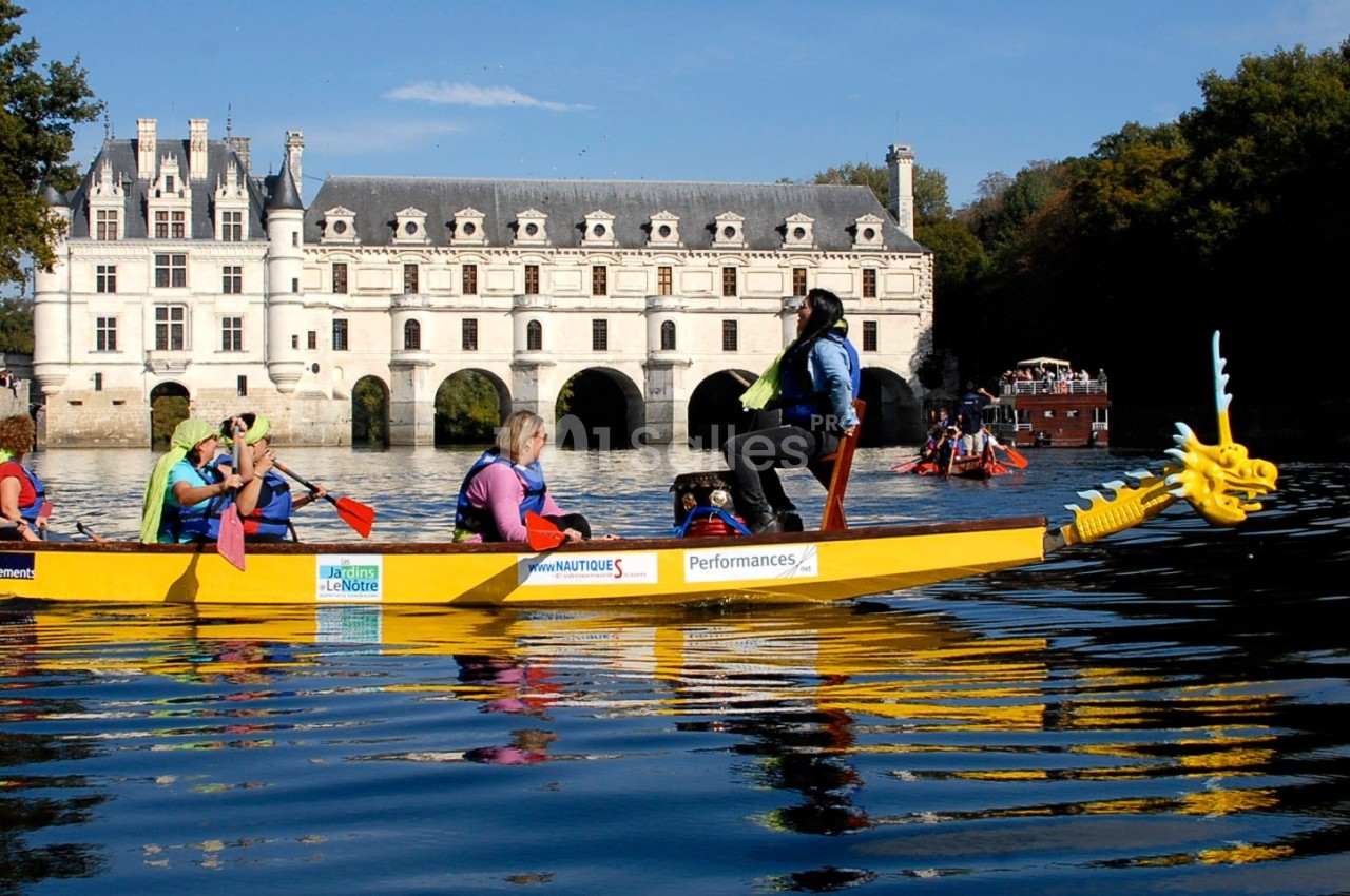Des personnes pagaient sur un bateau-dragon jaune devant le château de Chenonceau, entouré d'eau et de végétation.