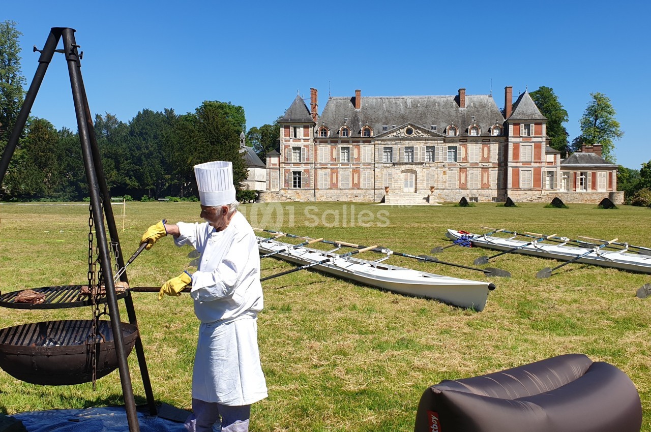 Un chef en tenue blanche cuisine au barbecue dans un parc près d'un château, avec des avirons posés sur l'herbe.