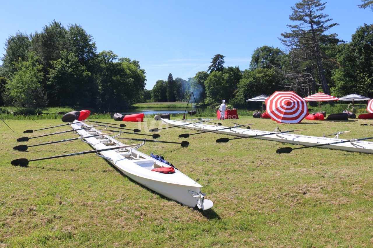 Deux avirons blancs posés sur une pelouse près d'un lac, entourés de parasols rouges et blancs et d'arbres.