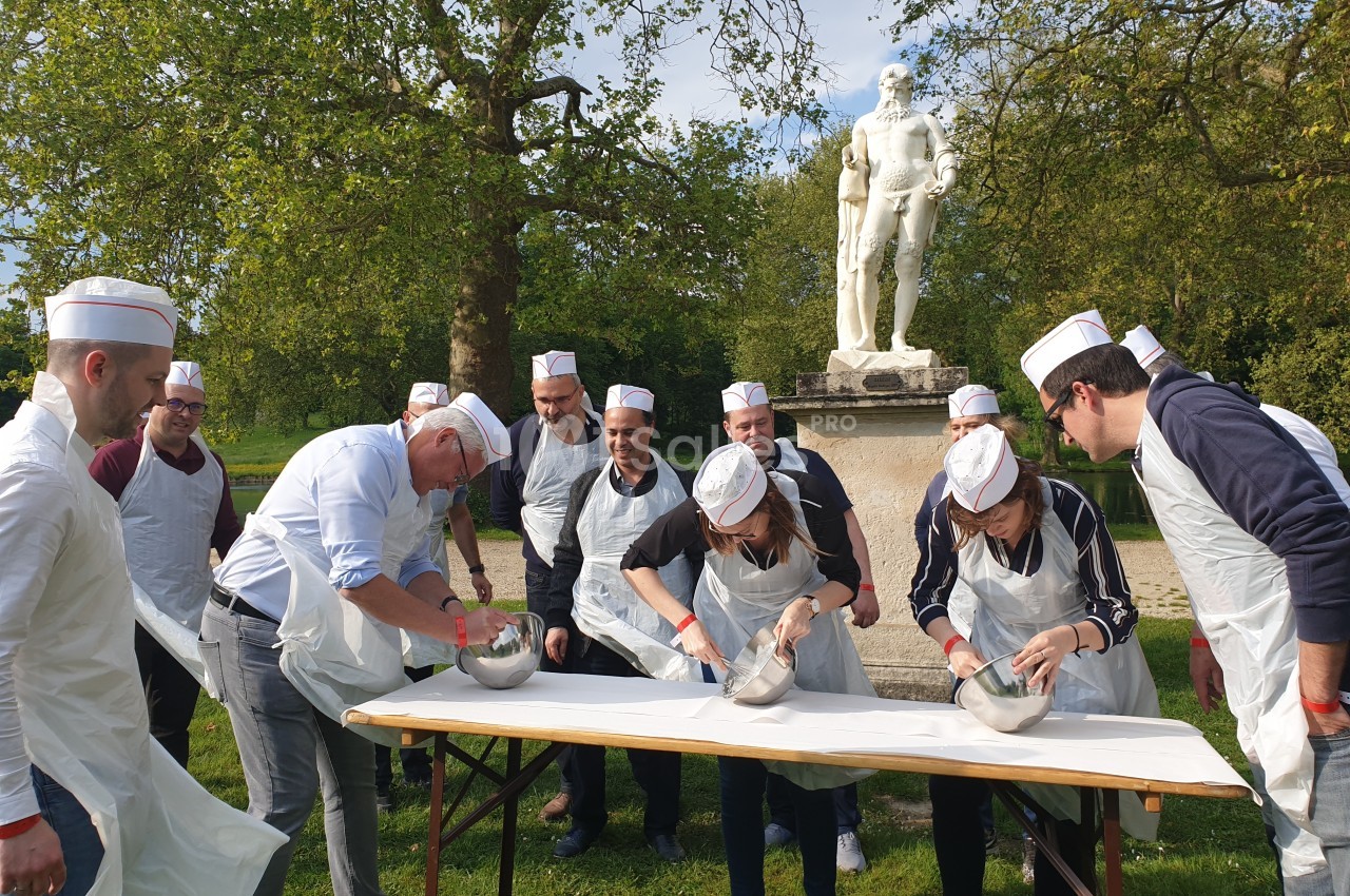 Des personnes en tabliers et toques participent à une activité culinaire en extérieur devant une statue dans un parc.