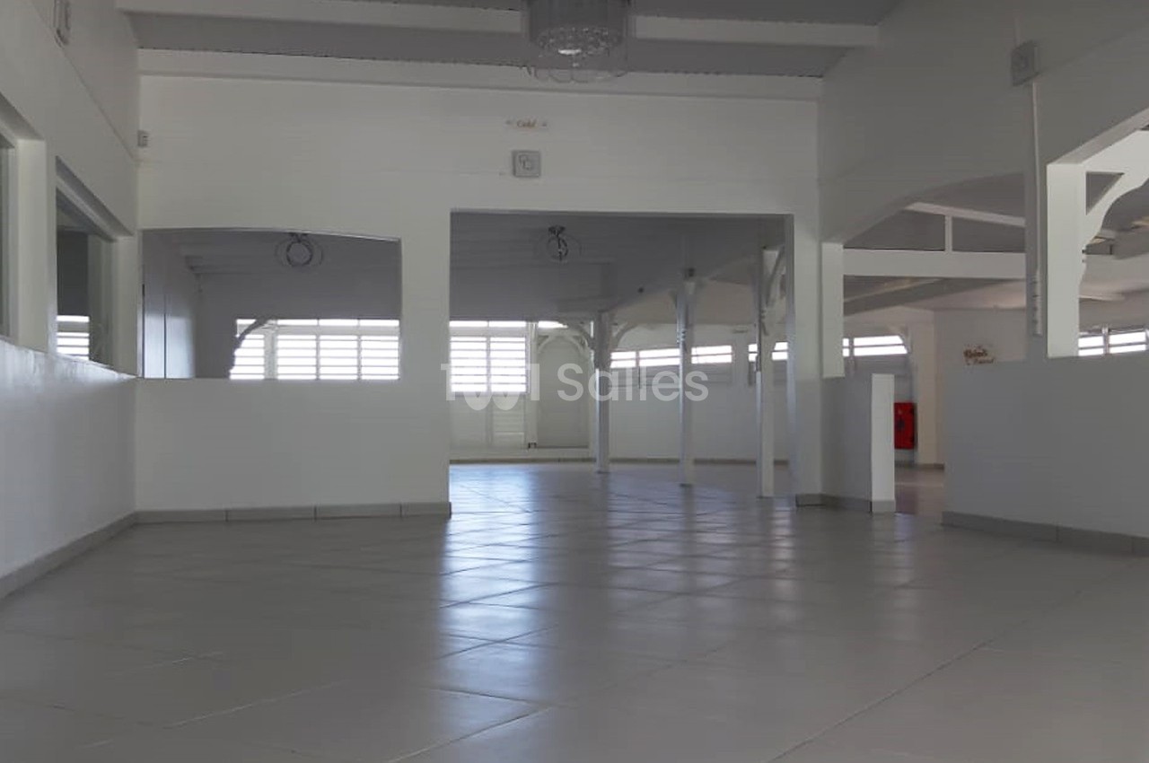 Salle vide avec sol carrelé, murs blancs, poutres apparentes et fenêtres laissant entrer la lumière naturelle.