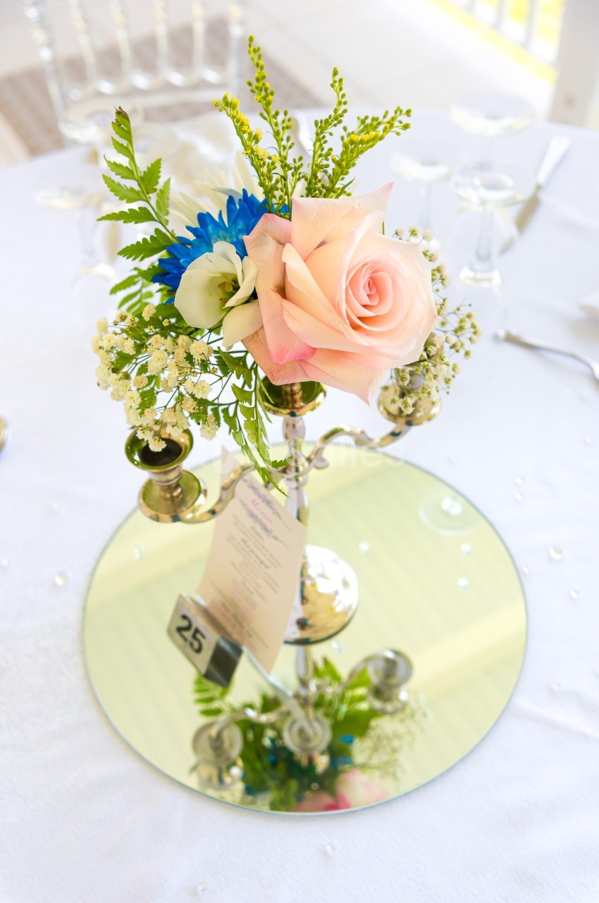 Centre de table floral avec une rose rose, des fleurs blanches et bleues, posé sur un miroir rond avec un numéro 25.