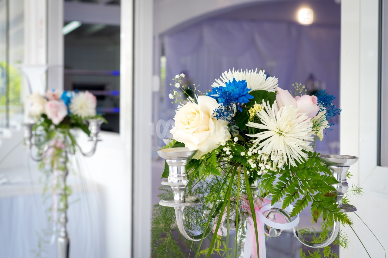 Arrangement floral coloré avec roses blanches, fleurs bleues et verdure, posé sur un chandelier argenté dans une salle…