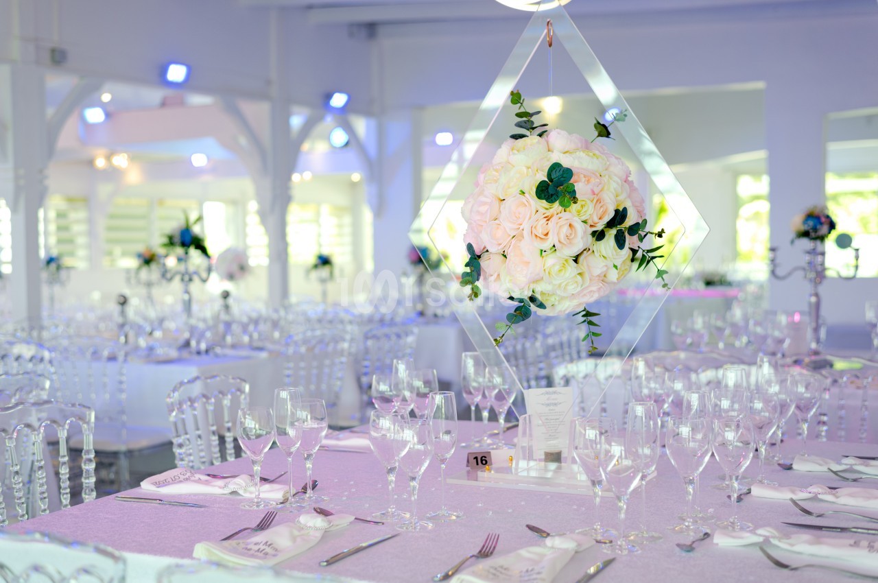 Salle de réception décorée avec des tables élégantes, nappes lilas, chaises transparentes et centre de table floral suspendu.