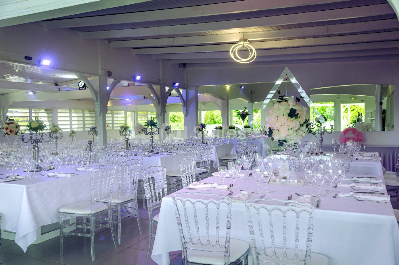 Salle de réception lumineuse avec tables dressées, chaises transparentes et décorations florales élégantes.