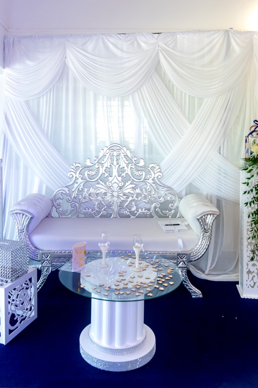 Canapé argenté décoratif devant un rideau blanc drapé, entouré de colonnes fleuries et d'une table basse en verre.
