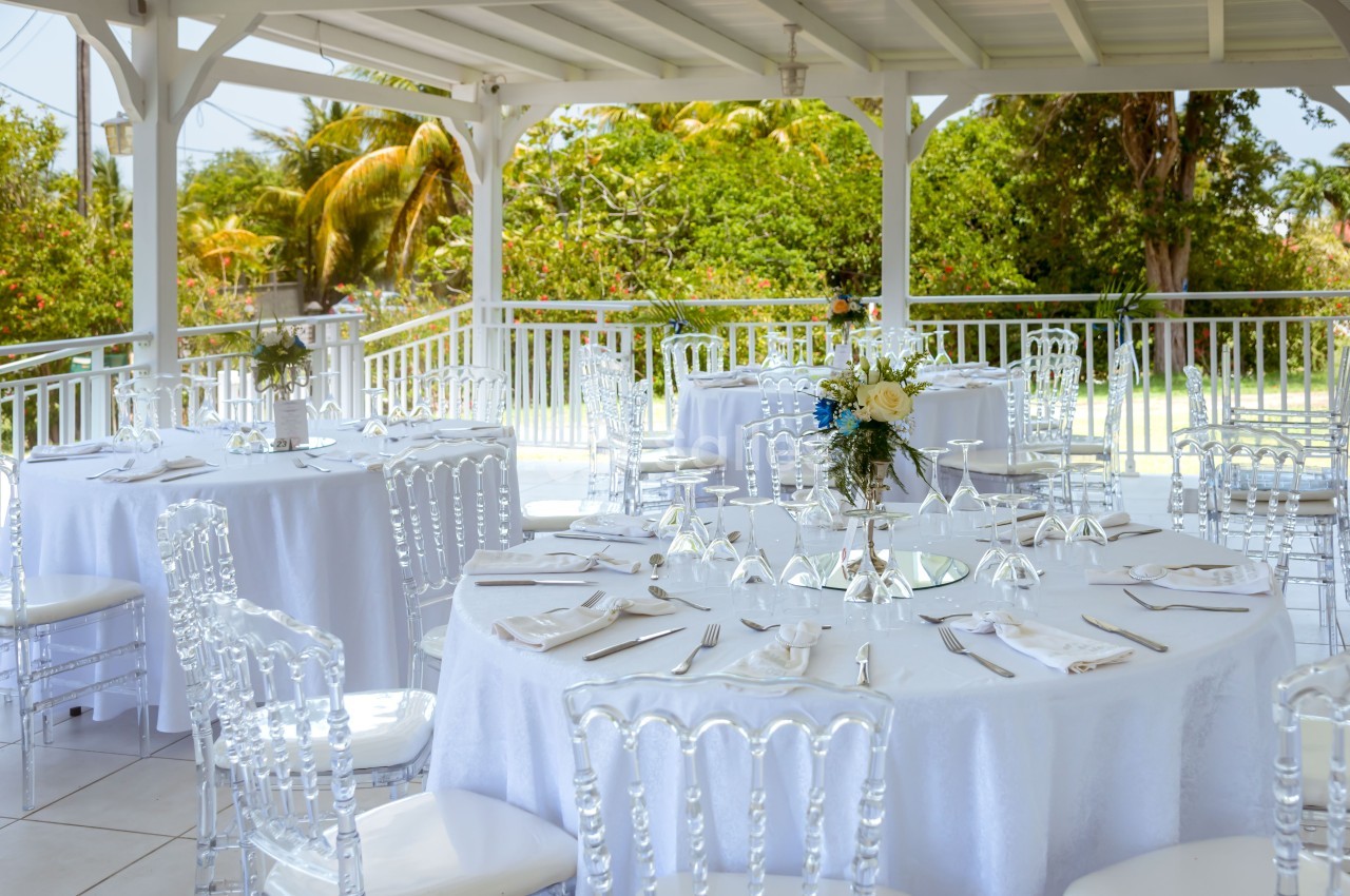 Tables rondes dressées avec nappes blanches et chaises transparentes sous une pergola, entourée de verdure.