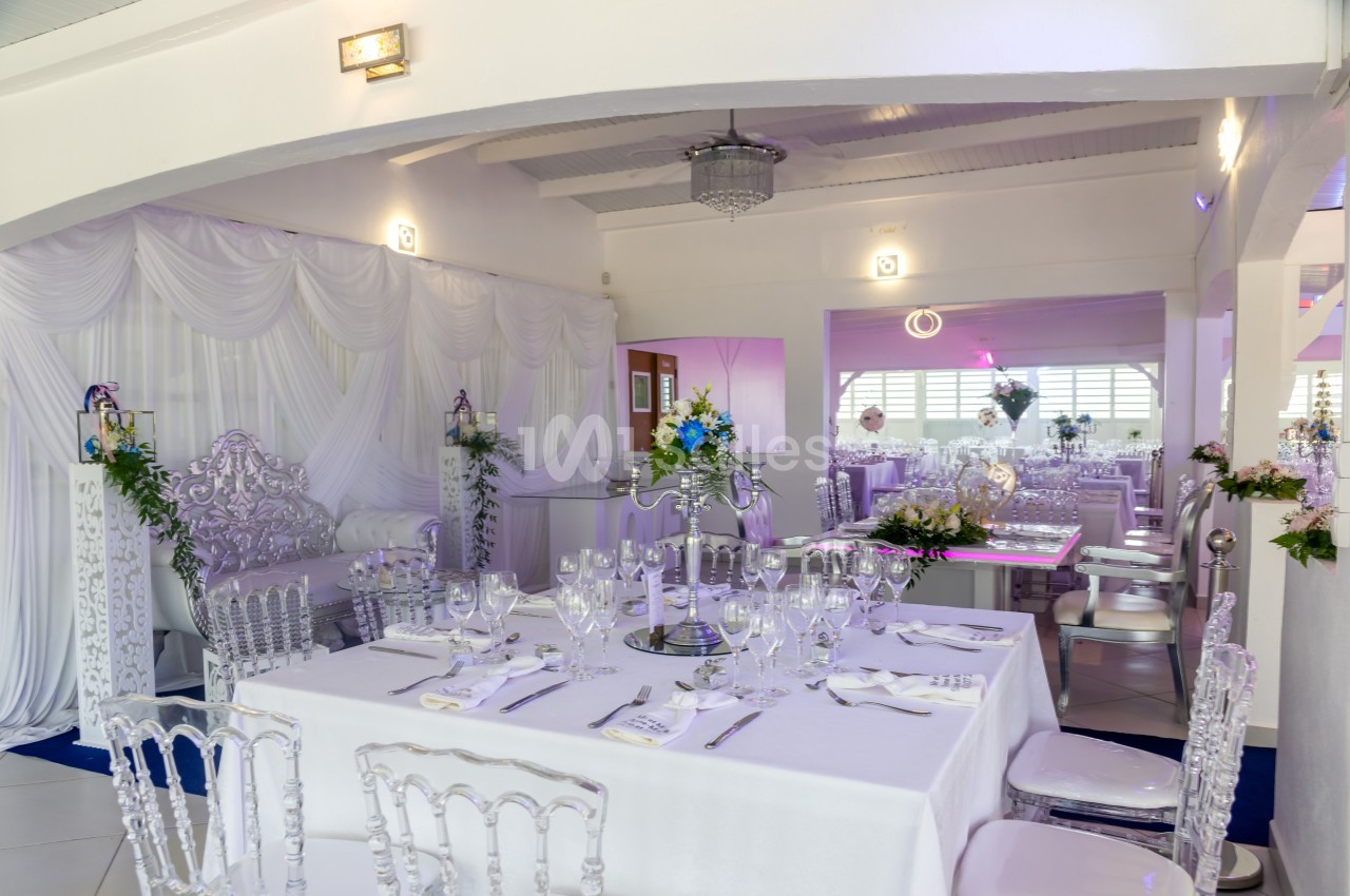 Salle de réception élégante avec tables dressées, nappes blanches, chaises transparentes et décorations florales.
