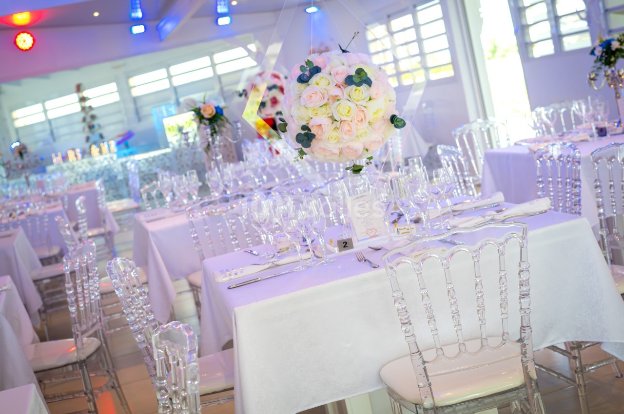 Salle de réception lumineuse avec tables dressées, nappes blanches, chaises transparentes et centre de table floral.