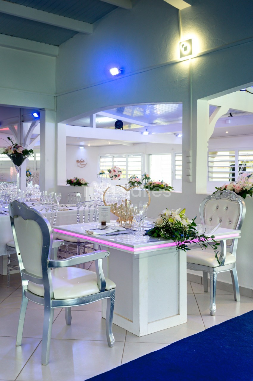 Table décorée avec des fleurs et des verres dans une salle lumineuse, avec des chaises argentées et un éclairage tamisé.
