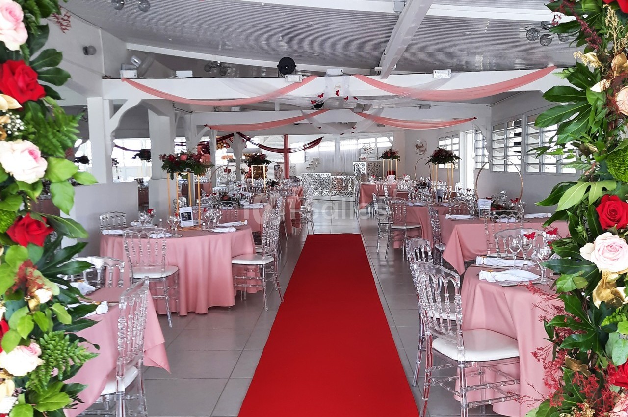Salle de réception décorée avec des tables roses, des chaises transparentes et un tapis rouge central.