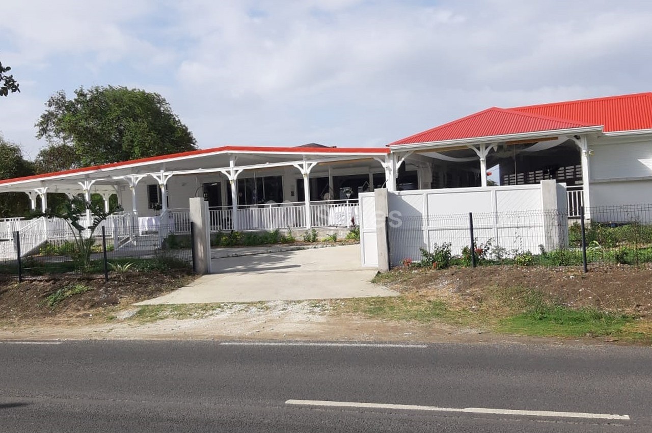 Bâtiment blanc avec toit rouge, entouré d'une clôture et d'un jardin, vu depuis une route asphaltée.