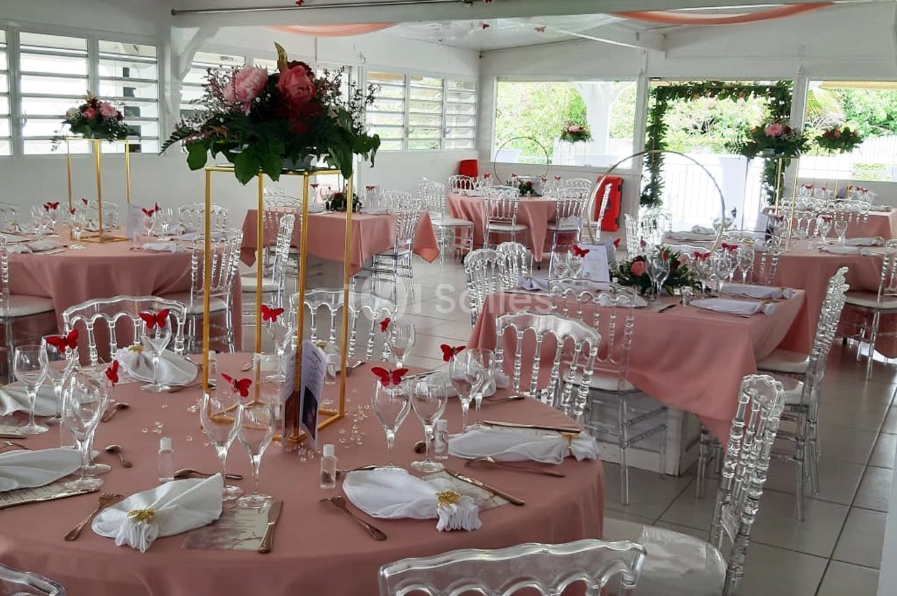 Salle de réception décorée avec des tables rondes, nappes roses, chaises transparentes et arrangements floraux.
