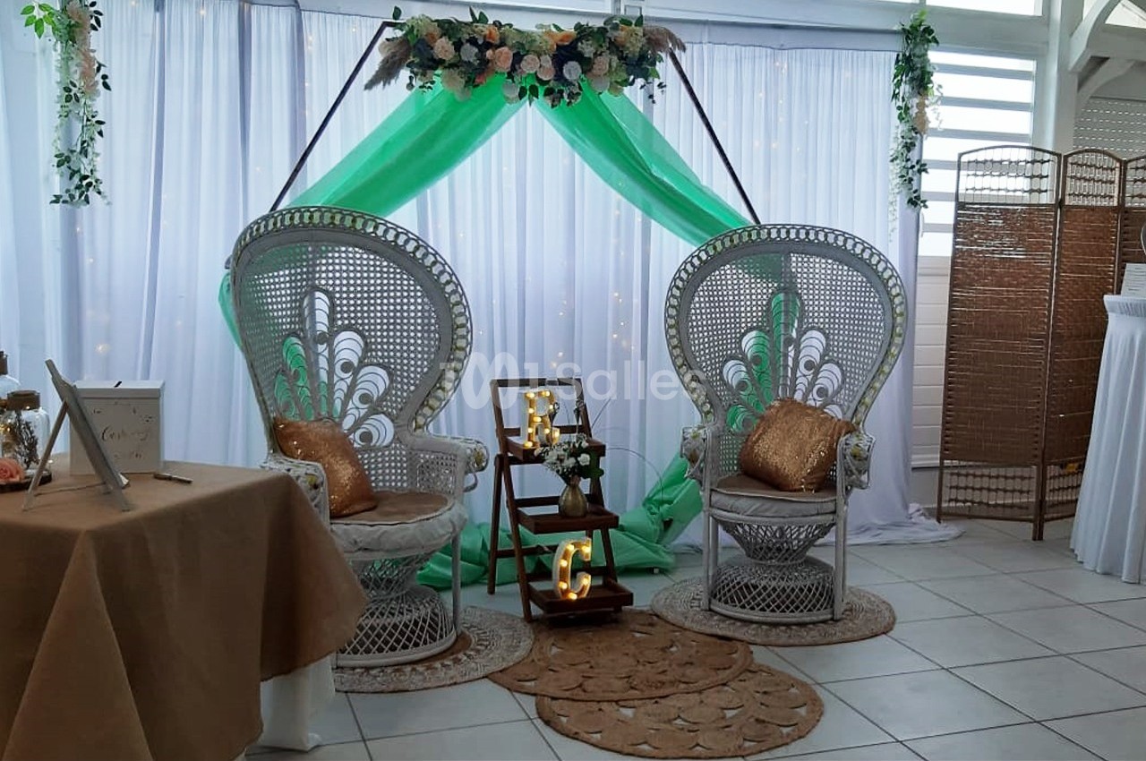 Deux fauteuils en rotin avec coussins dorés, décorés de fleurs et d'un drapé vert, dans un espace événementiel.