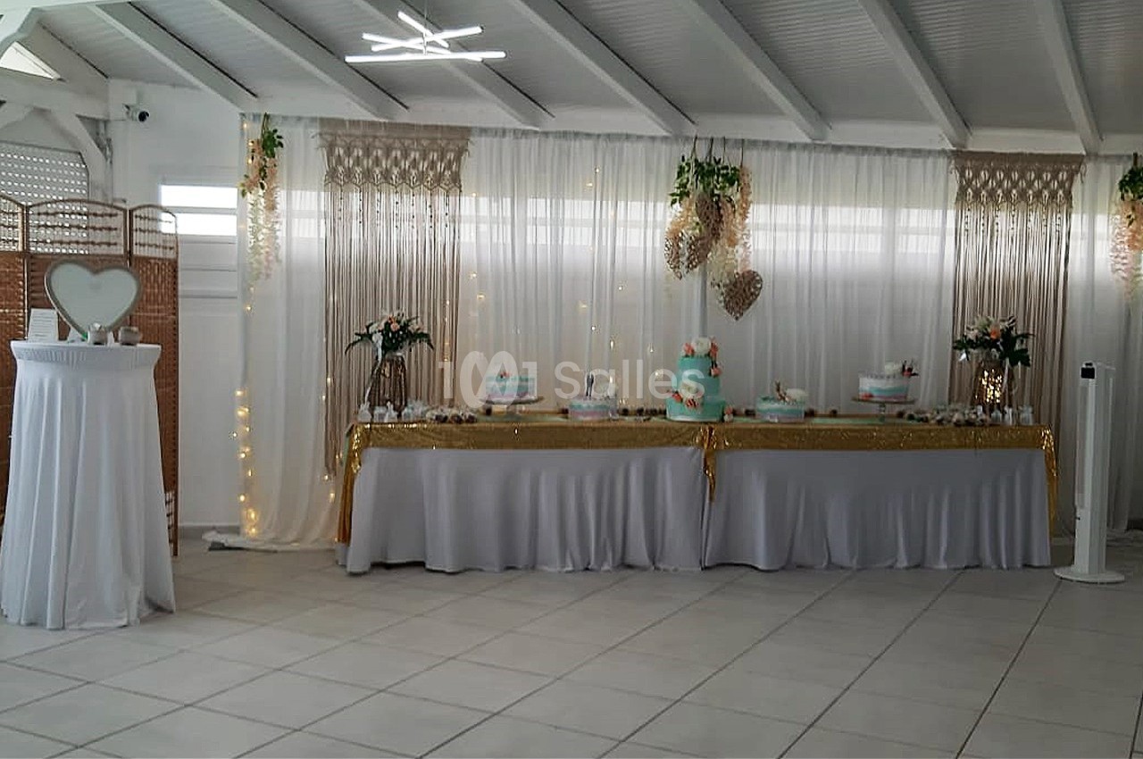 Table décorée pour une réception avec nappes blanches, gâteaux, fleurs et guirlandes lumineuses dans une salle lumineuse.