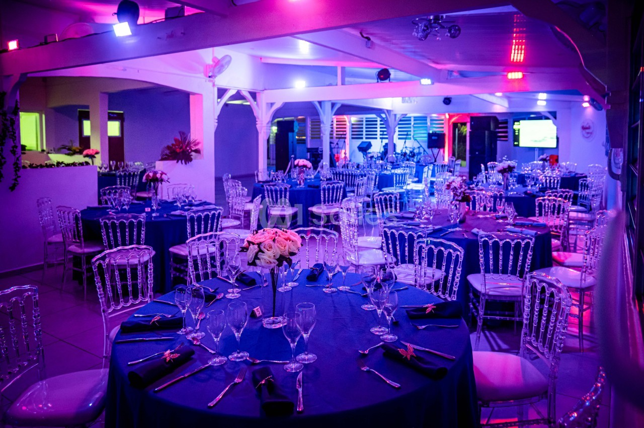 Salle de réception décorée avec des tables rondes dressées, éclairage coloré et ambiance festive.