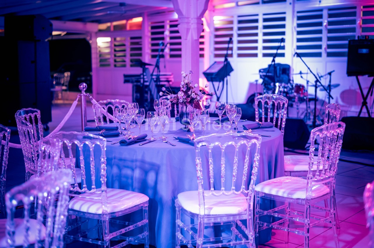 Salle de réception décorée avec des tables rondes, chaises transparentes et éclairage coloré, instruments en arrière-plan.
