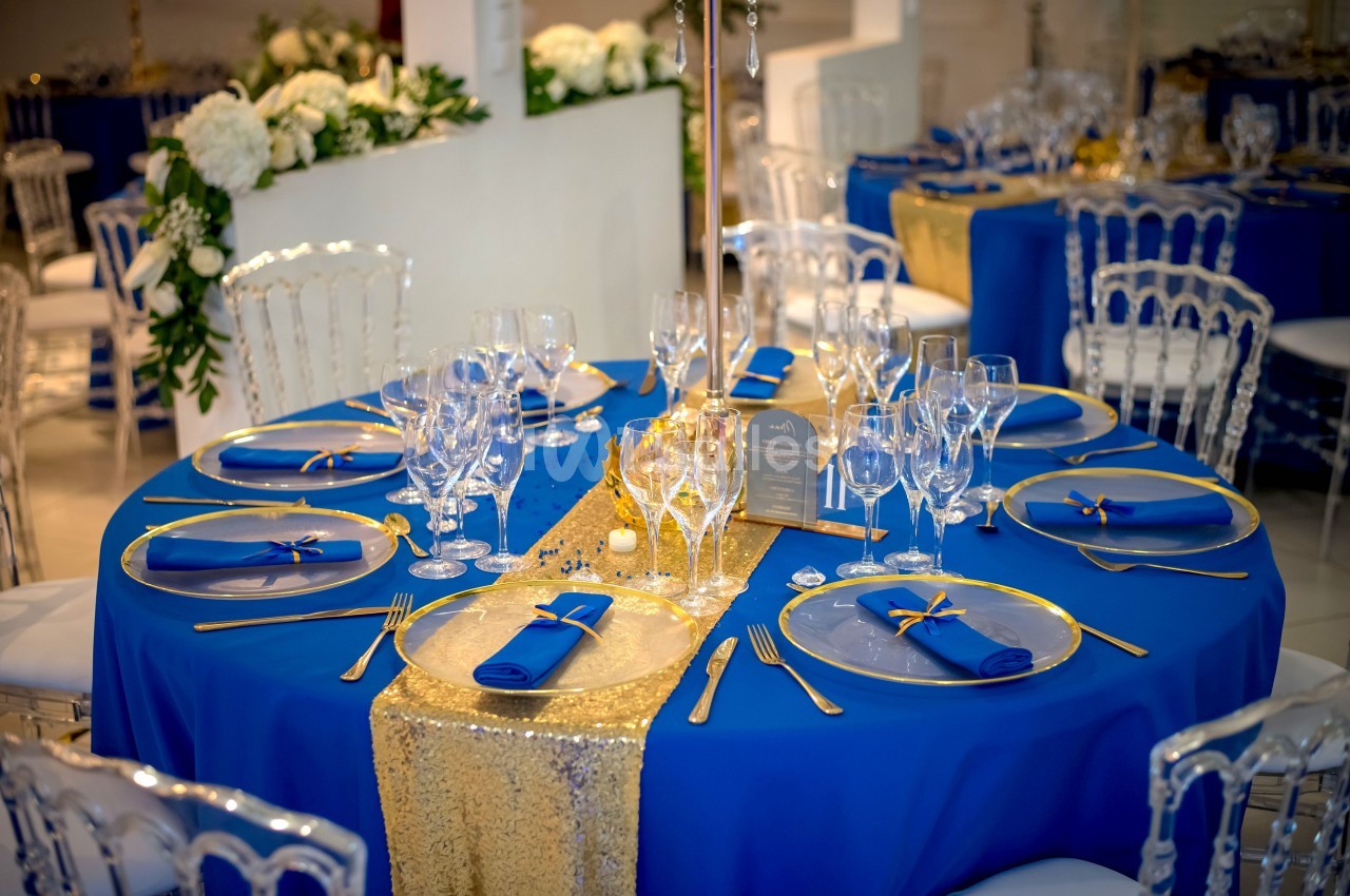 Table élégamment dressée avec nappes bleues, chemin de table doré, vaisselle raffinée et décorations florales blanches.