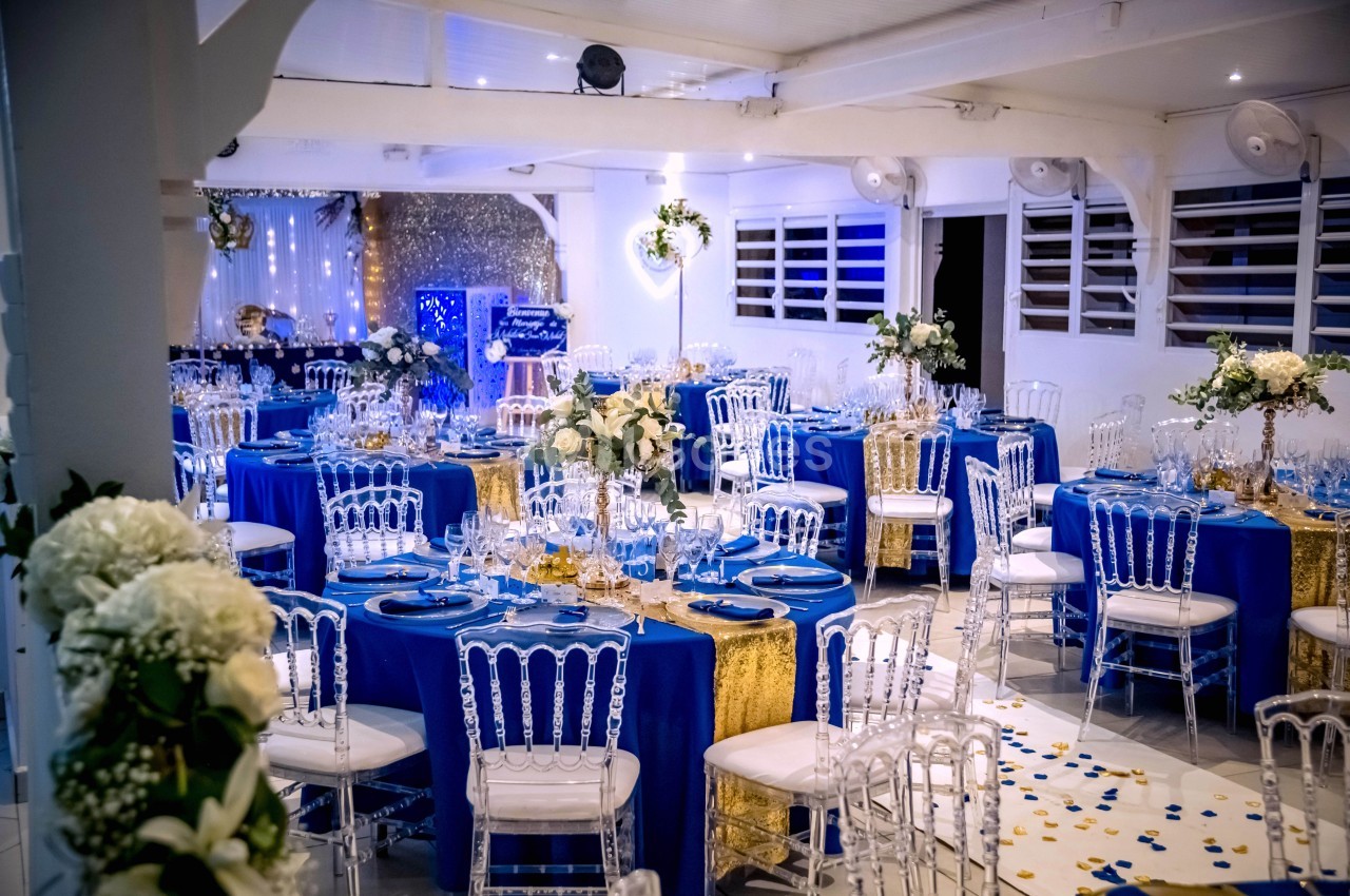 Salle de réception décorée avec des tables rondes, nappes bleues, chaises transparentes et arrangements floraux élégants.