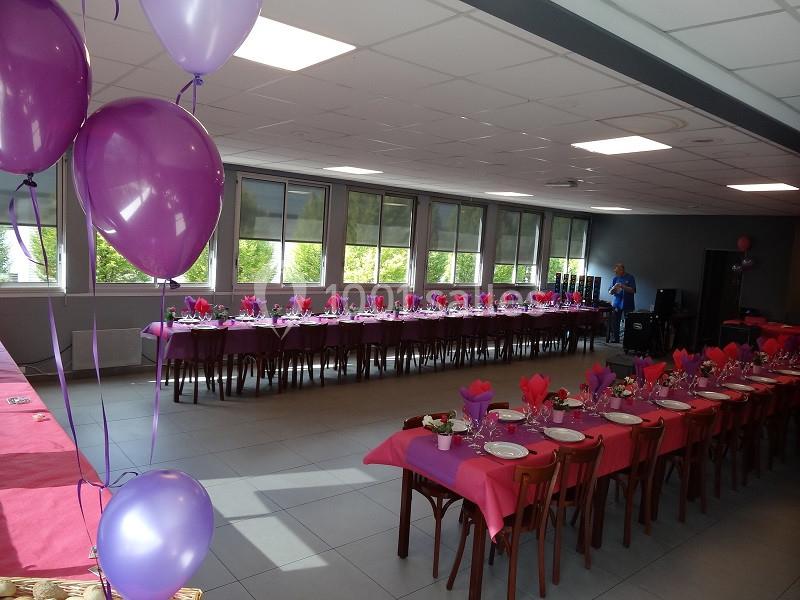 Salle décorée pour un événement avec tables dressées, nappes roses, serviettes pliées et ballons violets.