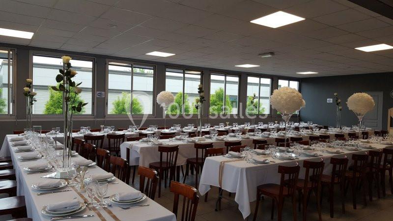 Salle de réception décorée avec des tables alignées, nappes blanches, vaisselle élégante et centres de table fleuris.
