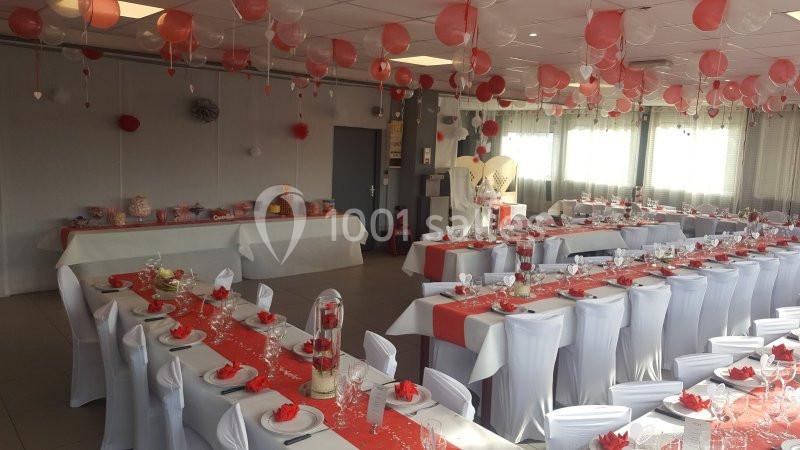 Salle décorée pour une réception avec tables dressées, nappes blanches et rouges, et ballons suspendus au plafond.
