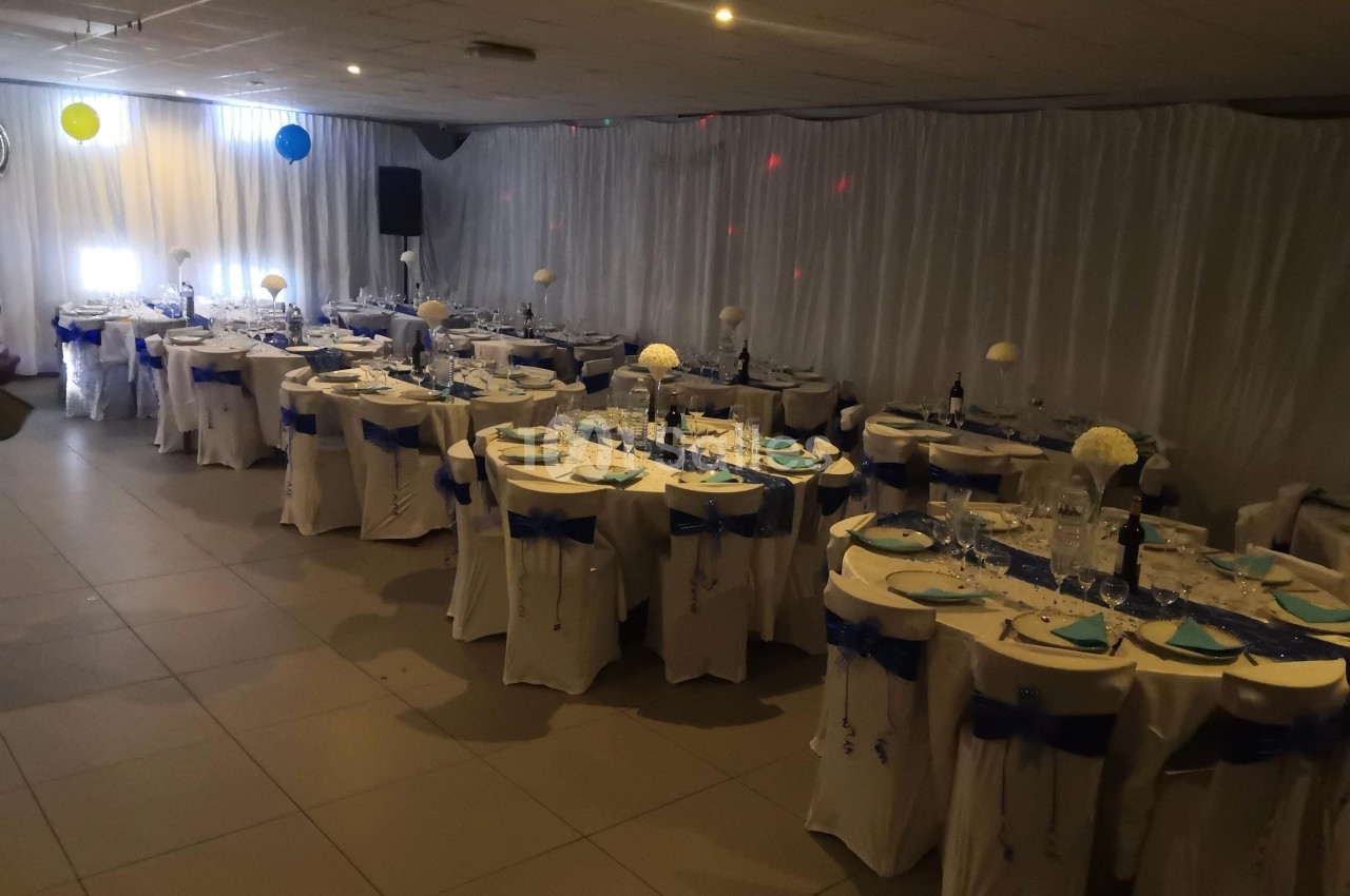 Salle de réception décorée avec des tables rondes dressées, nappes blanches et rubans bleus, éclairage tamisé.