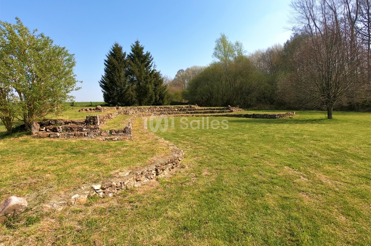 Ruines en pierre dans un espace vert dégagé, entouré d'arbres et sous un ciel bleu clair.