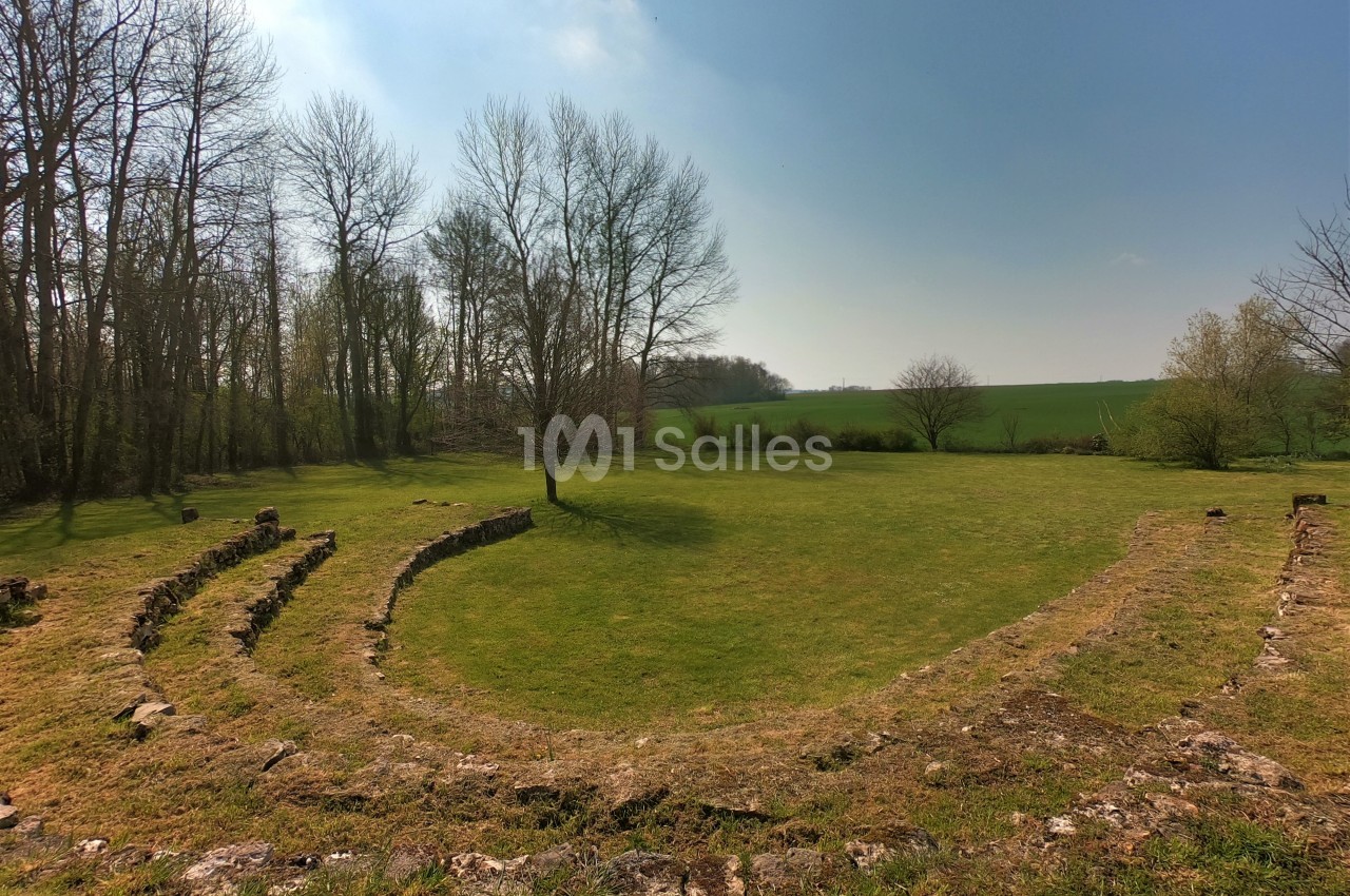 Vestiges d'un petit amphithéâtre en pierre entouré d'arbres et donnant sur un champ verdoyant sous un ciel dégagé.