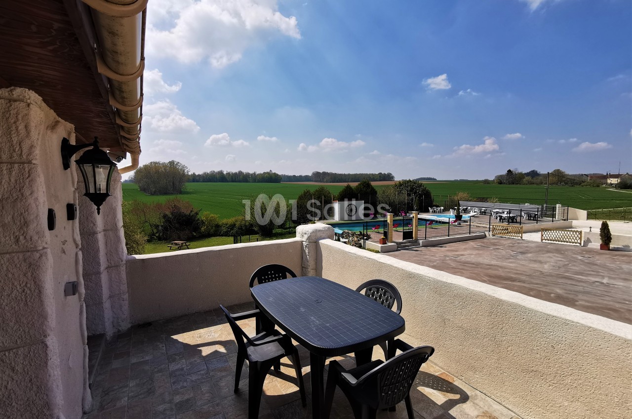 Terrasse avec table et chaises noires, vue sur une piscine, un jardin et des champs sous un ciel partiellement nuageux.