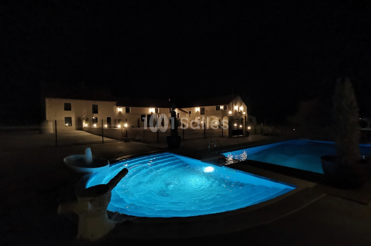Vue nocturne d'une piscine éclairée en premier plan, avec un bâtiment illuminé en arrière-plan.