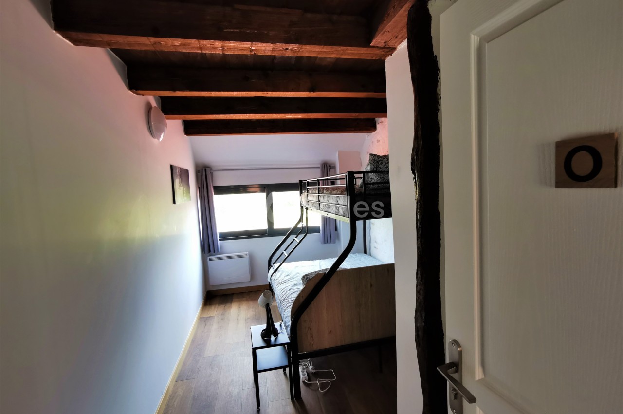 Chambre avec lit superposé en bois, parquet au sol, poutres apparentes et grande fenêtre laissant entrer la lumière.
