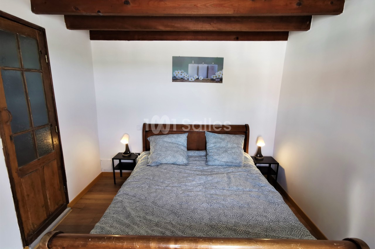 Lit double en bois avec une couverture bleue, encadré par deux lampes de chevet, dans une chambre aux poutres apparentes.