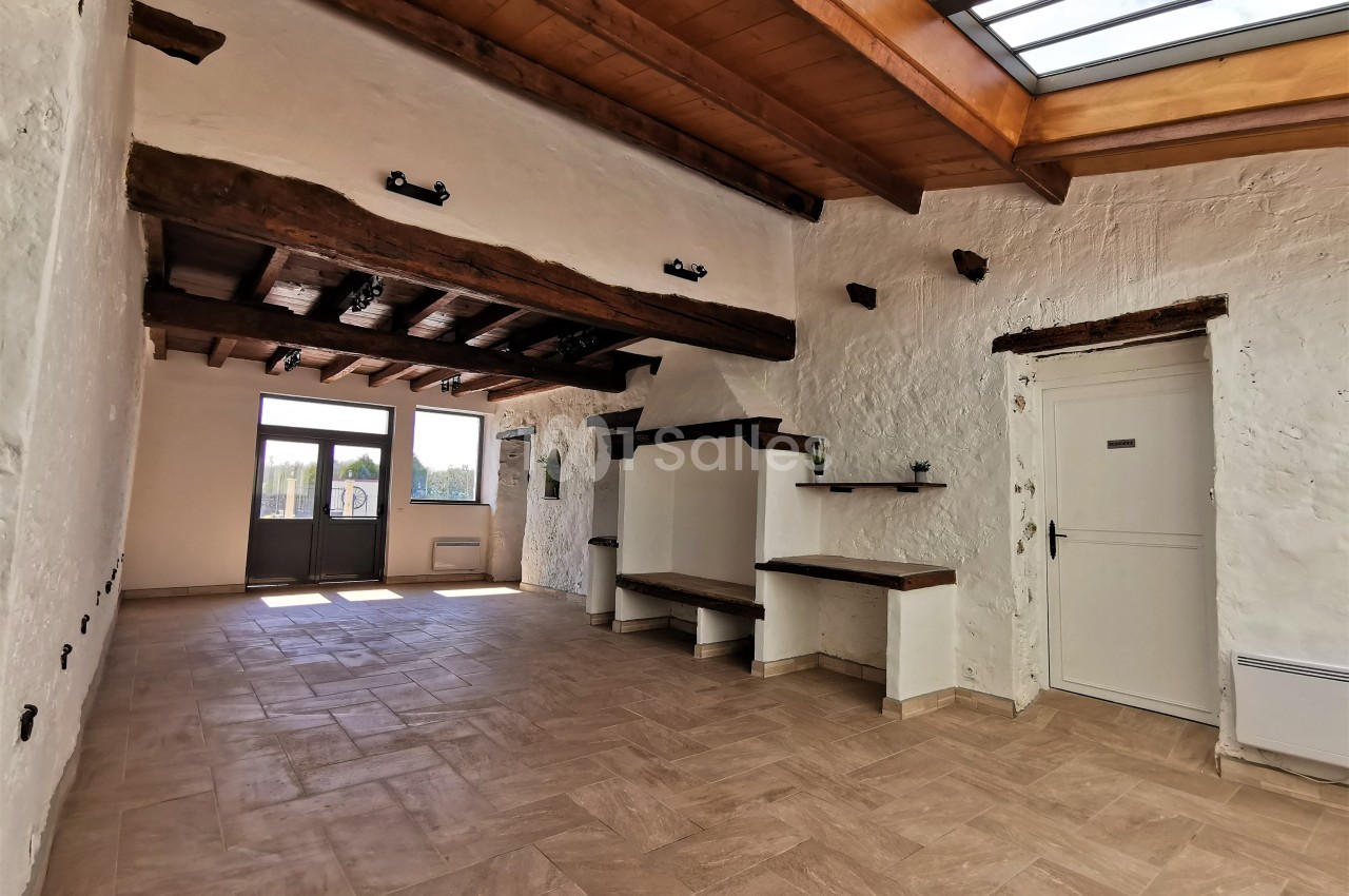 Salle lumineuse avec murs en pierre blanche, poutres apparentes, sol en carrelage beige et grande verrière au plafond.