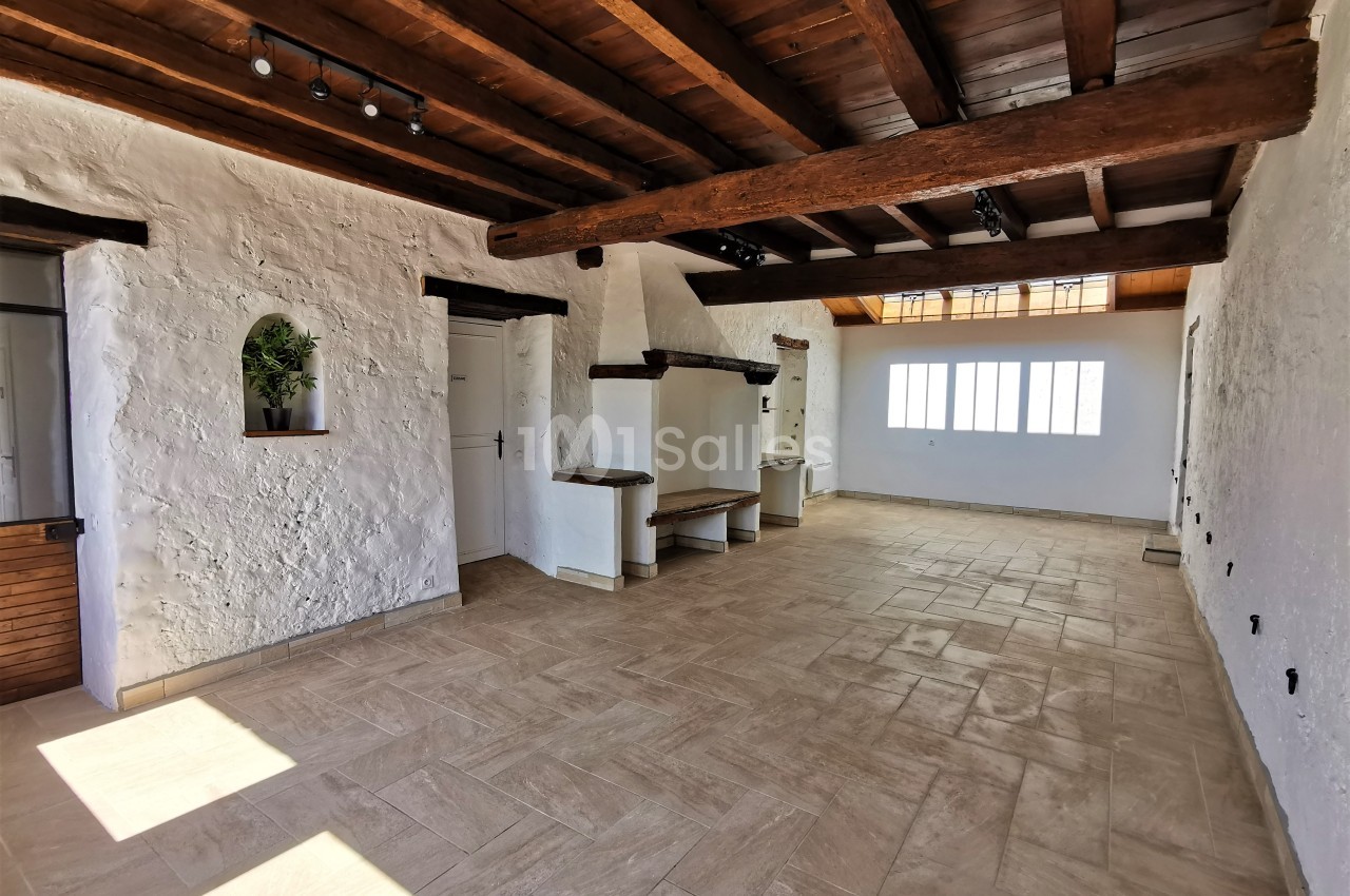 Salle lumineuse avec murs blancs texturés, poutres en bois au plafond, sol en carrelage beige et grandes fenêtres.