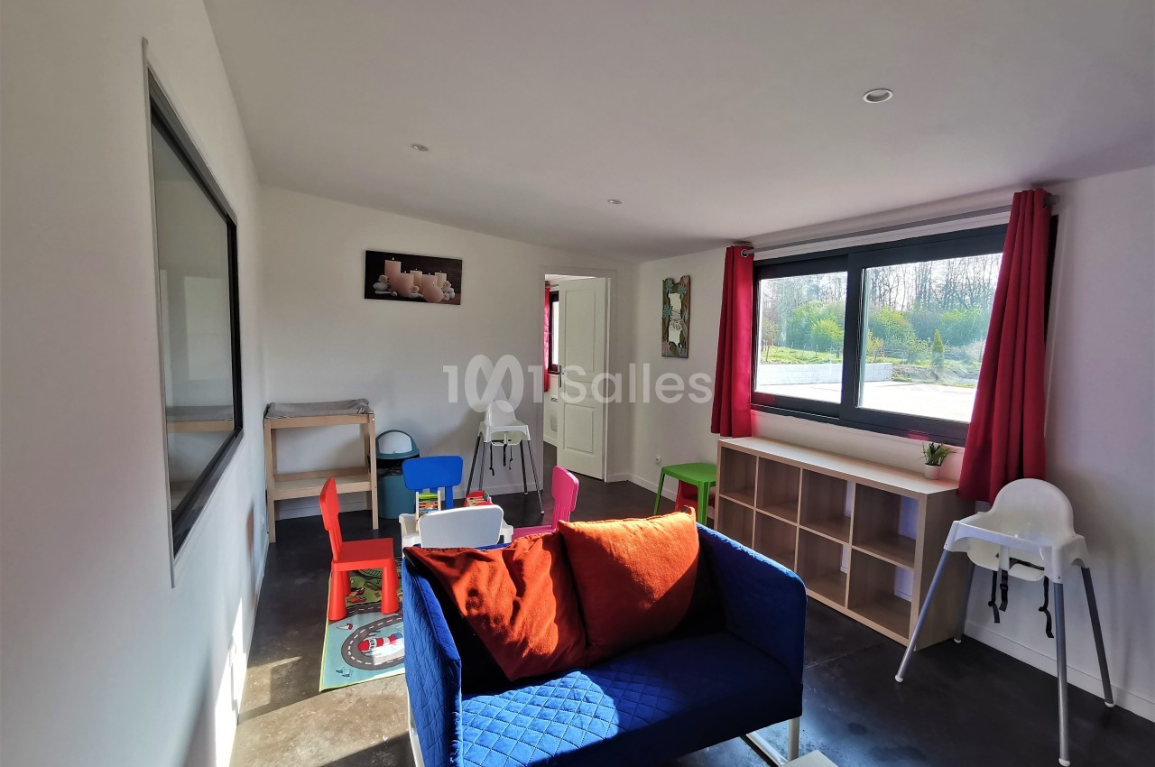 Salle lumineuse avec canapé bleu, chaises colorées, jouets pour enfants, et grande fenêtre donnant sur un extérieur…