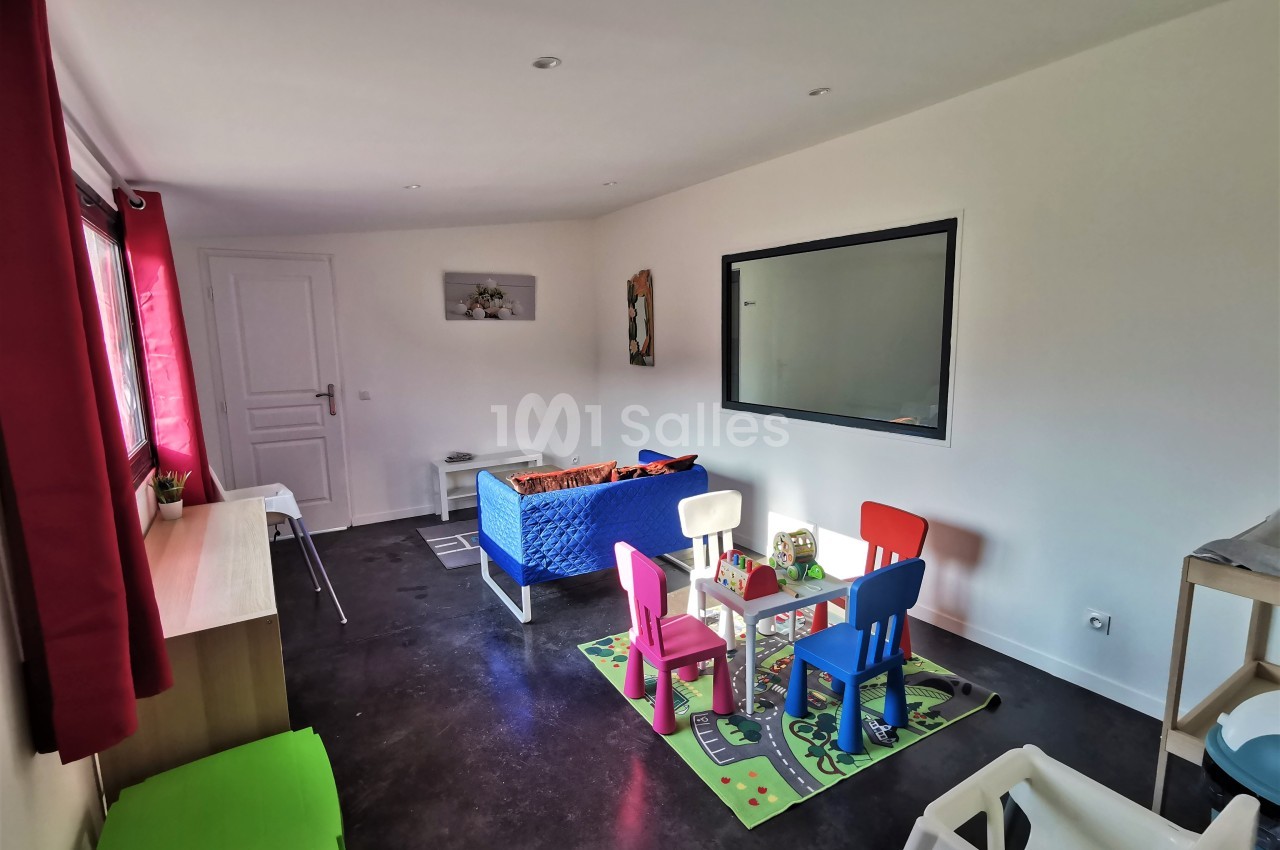 Salle de jeux lumineuse avec tapis coloré, petites chaises, table et lit pour enfants, murs blancs et fenêtre.