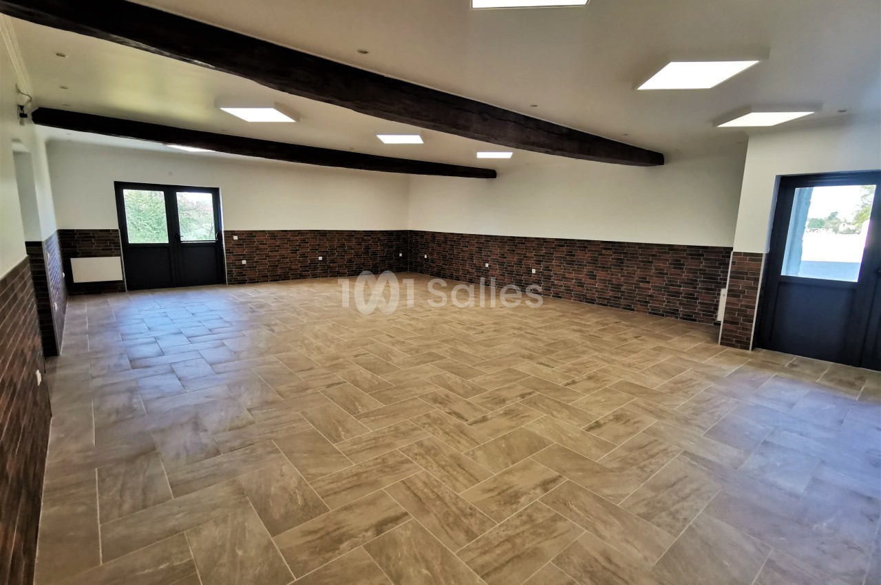 Salle vide avec carrelage beige, murs partiellement en briques, poutres apparentes et grandes fenêtres laissant entrer la…