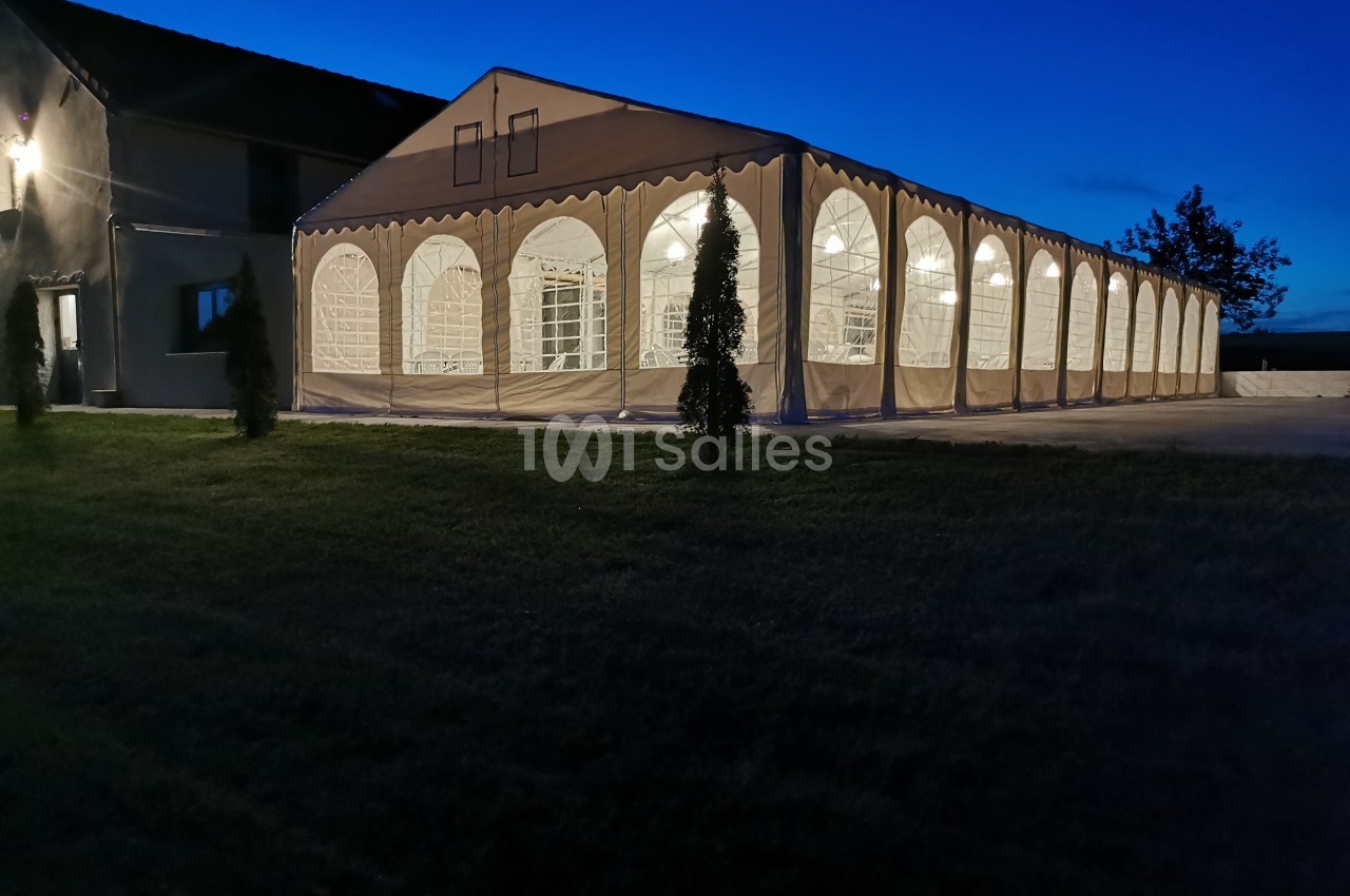 Grande tente éclairée de nuit, installée sur une surface pavée à côté d'un bâtiment.