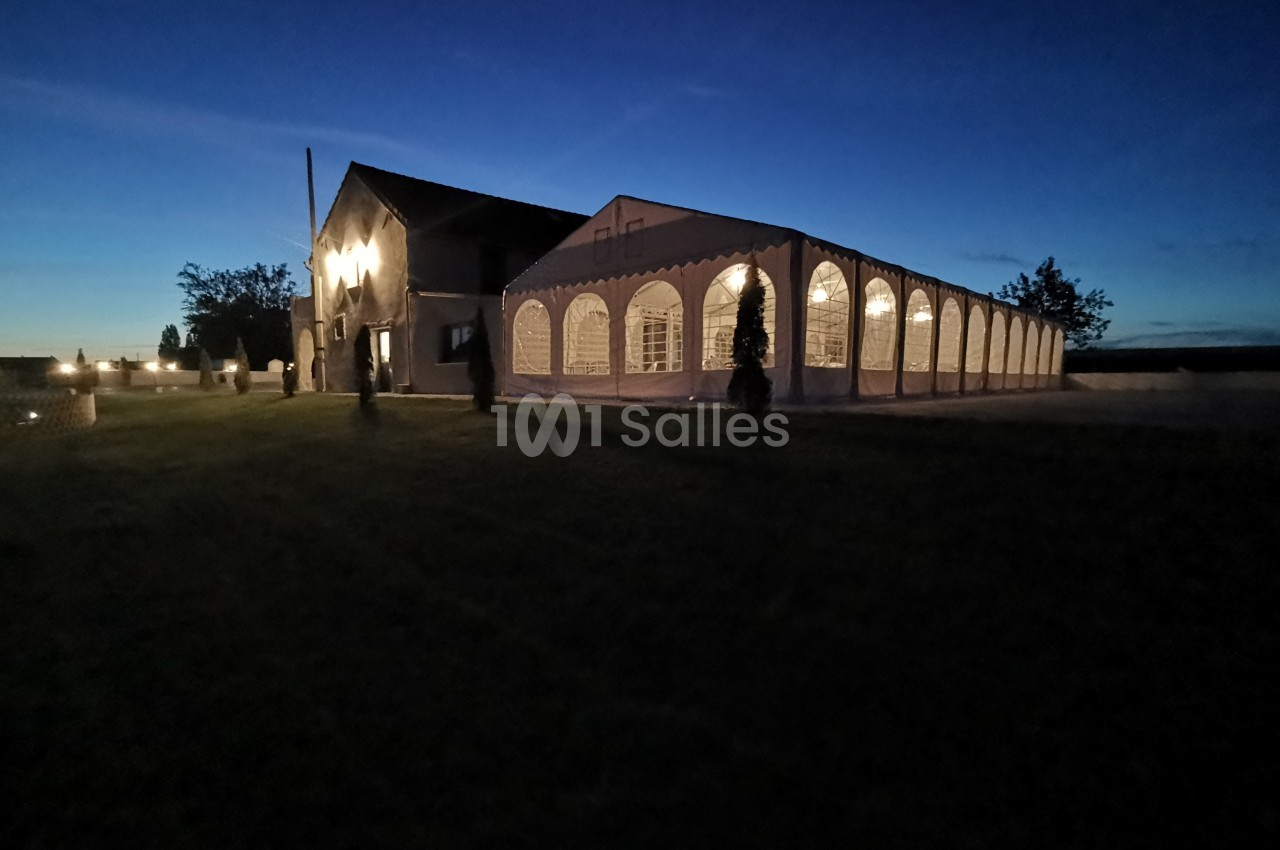 Grande tente éclairée installée près d'un bâtiment en pierre, dans un paysage nocturne avec ciel bleu foncé.