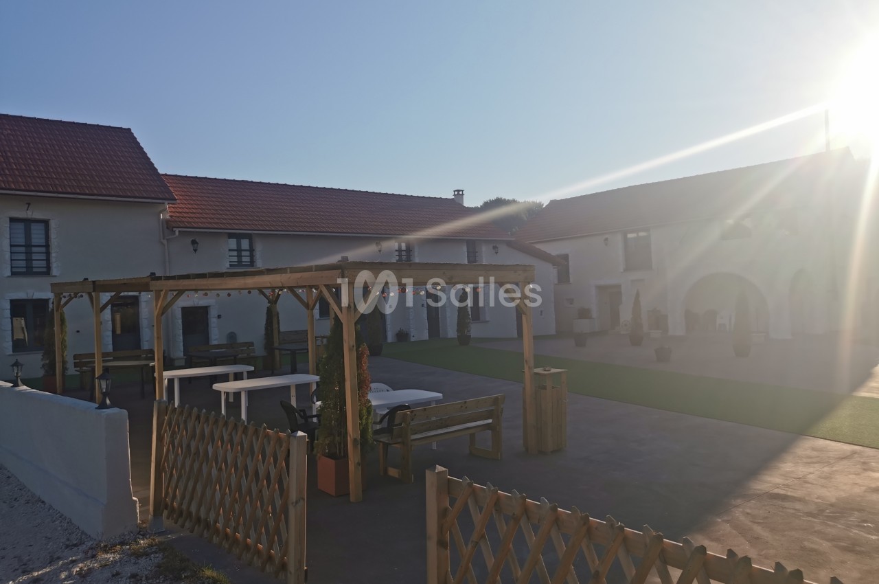 Cour extérieure avec pergola en bois, tables et bancs, entourée de bâtiments aux toits rouges sous un soleil éclatant.