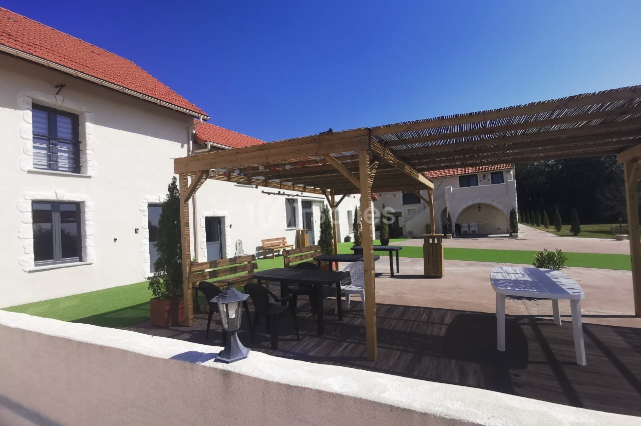 Pergola en bois avec tables et bancs sur une terrasse ensoleillée, entourée de bâtiments blancs avec toits rouges.