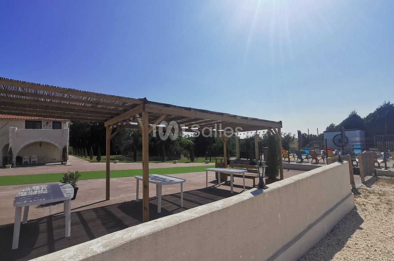 Terrasse extérieure avec pergola en bois, tables blanches, pelouse et piscine en arrière-plan sous un ciel ensoleillé.
