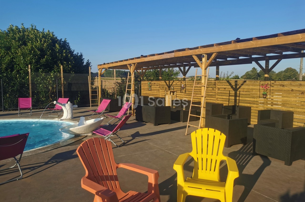 Terrasse ensoleillée avec piscine, chaises colorées, transats et pergola en bois dans un cadre extérieur calme.